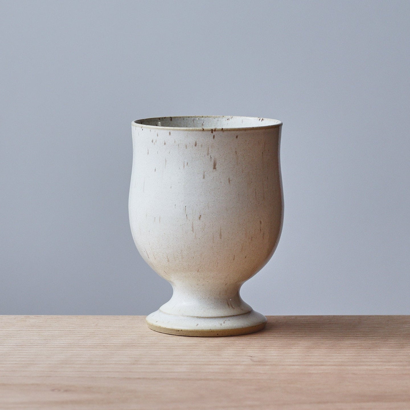 A small ceramic cup with a speckled Ivory White glaze, sitting on a light wood surface against a solid gray background.