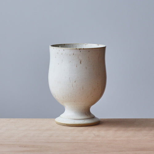 A small ceramic cup with a speckled Ivory White glaze, sitting on a light wood surface against a solid gray background.