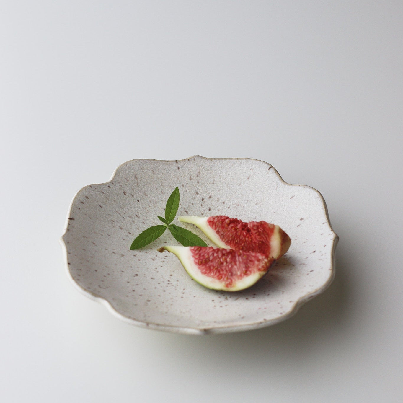 A small scalloped ceramic plate with a speckled off-white glaze, styled with a sliced fig and a green leaf on a clean light gray background.
