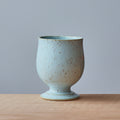 A small ceramic cup with a flared pedestal base, featuring a speckled light blue and beige glaze, set on a light wood surface against a solid gray background.