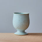 A small ceramic cup with a flared pedestal base, featuring a speckled light blue and beige glaze, set on a light wood surface against a solid gray background.