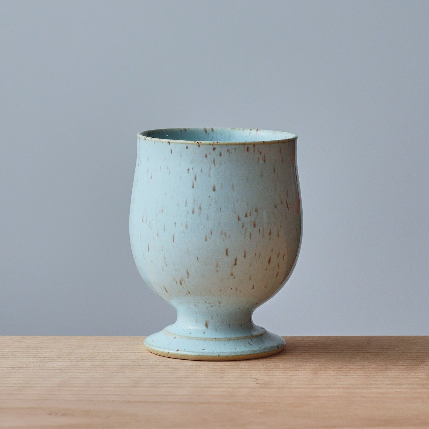 A small ceramic cup with a flared pedestal base, featuring a speckled light blue and beige glaze, set on a light wood surface against a solid gray background.