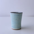A small handmade ceramic cup with a light blue speckled glaze and a dark rim, standing on a white surface against a light gray background.