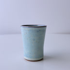 A small handmade ceramic cup with a light blue speckled glaze and a dark rim, standing on a white surface against a light gray background.