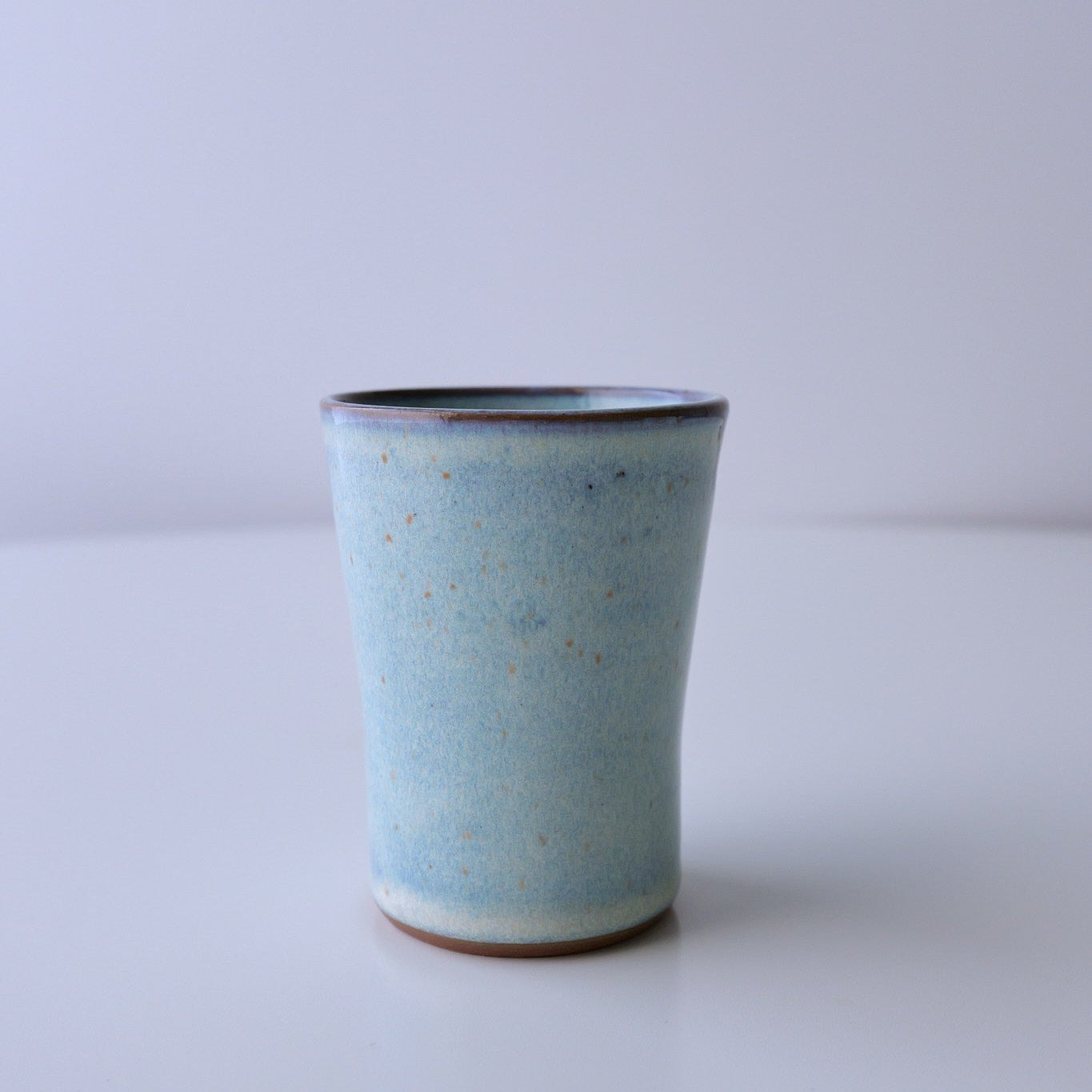 A small handmade ceramic cup with a light blue speckled glaze and a dark rim, standing on a white surface against a light gray background.