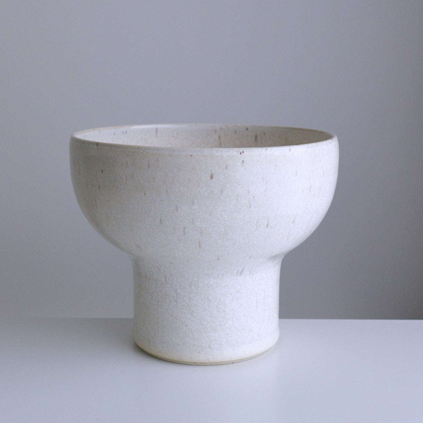A handmade ceramic bowl with a textured, speckled off-white glaze, standing on a matching cylindrical pedestal base against a neutral gray background.