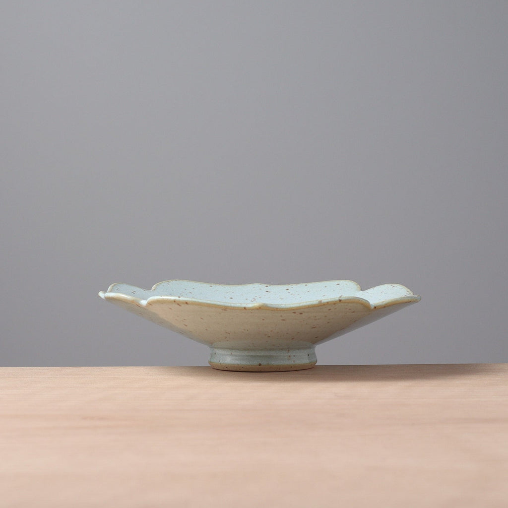 A small ceramic dish with a scalloped rim and a light blue-green glaze, sitting on a light wood surface against a solid gray background.
