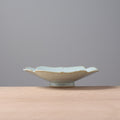 A small ceramic dish with a scalloped rim and a light blue-green glaze, sitting on a light wood surface against a solid gray background.