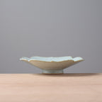 A small ceramic dish with a scalloped rim and a light blue-green glaze, sitting on a light wood surface against a solid gray background.