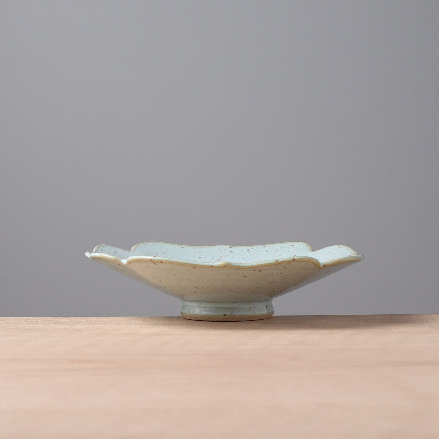 A small ceramic dish with a scalloped rim and a light blue-green glaze, sitting on a light wood surface against a solid gray background.
