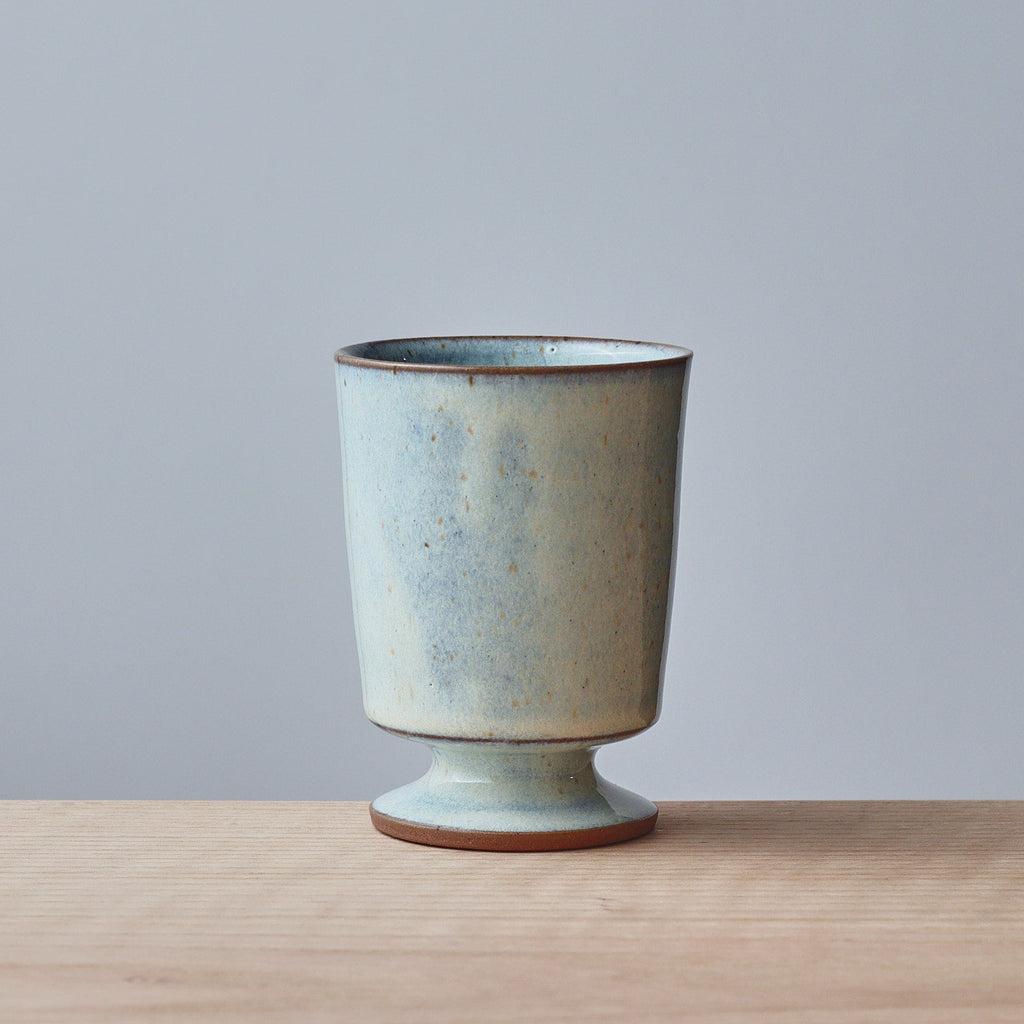 A small ceramic cup/goblet with a flared pedestal base, featuring a speckled light floating blue glaze, set on a light wood surface against a solid gray background.
