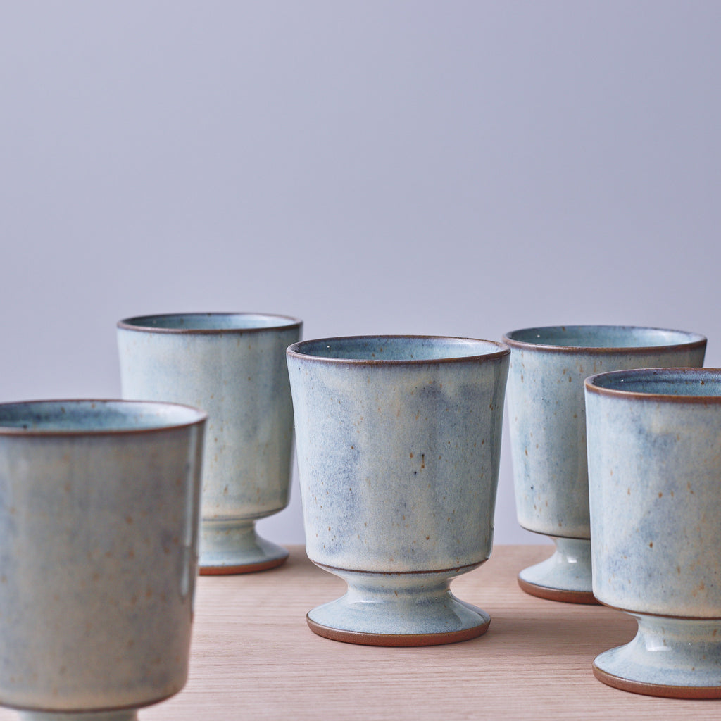 A set of five handmade ceramic cups with a light blue-green glaze and a brown rim, arranged on a light wooden surface against a neutral gray background.