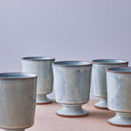 A set of five handmade ceramic cups with a light blue-green glaze and a brown rim, arranged on a light wooden surface against a neutral gray background.